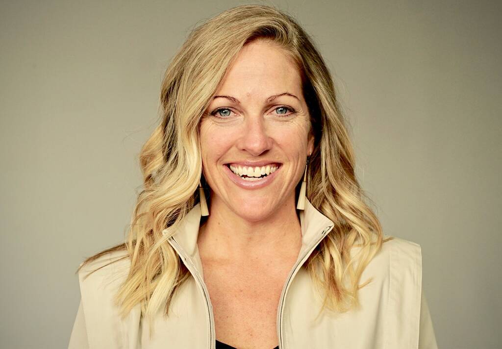 Headshot of a Nicole Noren smiling. She has shoulder-length blonde hair, is wearing a light beige vest and matching earrings against a neutral gray background.