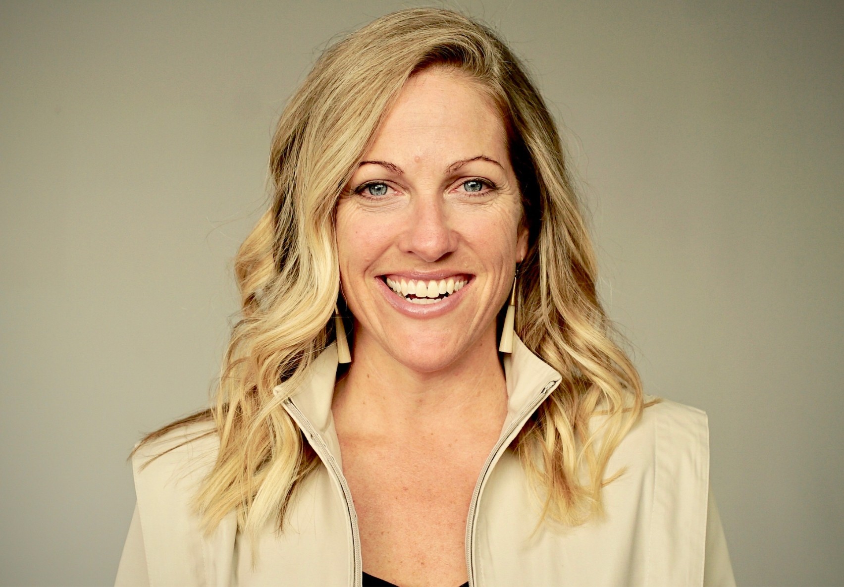 Headshot of a Nicole Noren smiling. She has shoulder-length blonde hair, is wearing a light beige vest and matching earrings against a neutral gray background.